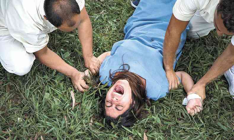 vestindo uniforme azul, atriz Carol Castro grita e e é contida pelos pulsos, deitada no chão, em cena de Insânia
