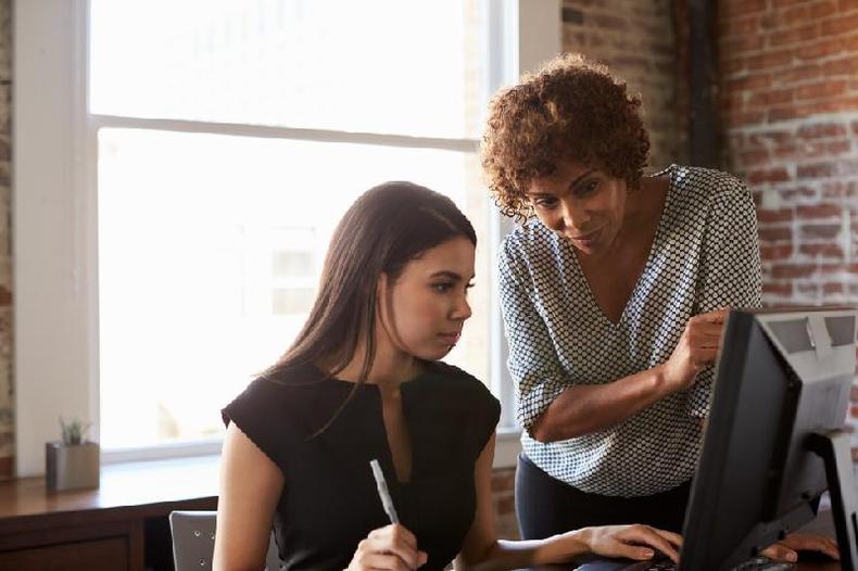 Segundo West Duffy, no processo de job crafting, é importante ter em mente as razões e motivações do nosso trabalho(foto: Getty Images) mulher branca sentada em frente a um computador conversa com mulher negra de pé