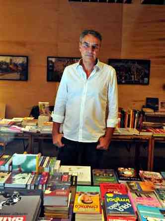 O jornalista Jos Eduardo Gonalves, em p, atrs de mesa cheia de livros, olha para a cmera 