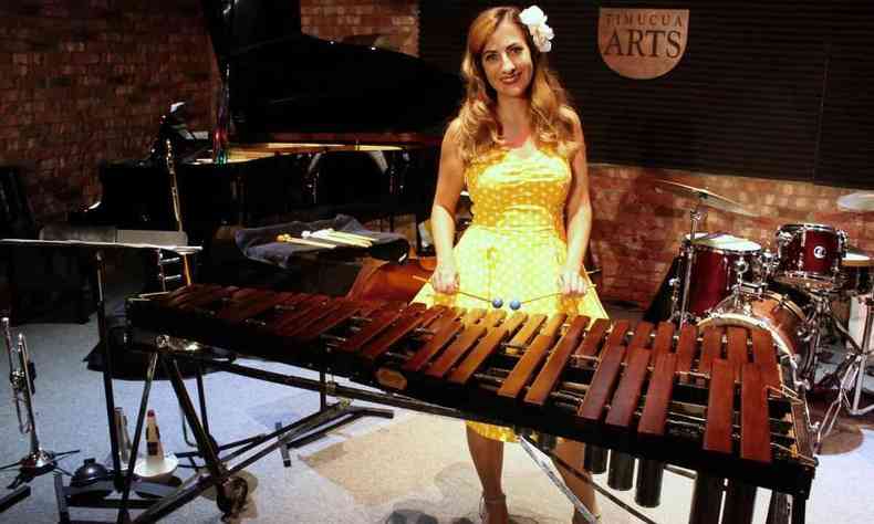 De vestido amarelo, Heather Thorn, em frente ao xilofone, olha para a câmera e sorri