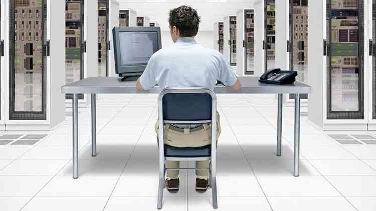 Homem sentado em frente a computador com salas repletas de equipamentos