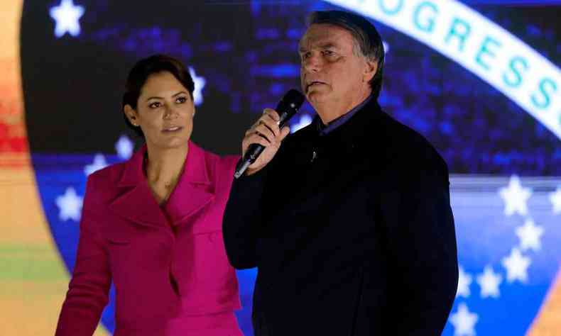 Michelle Bolsonaro olha para o marido, o presidente Jair Bolsonaro, que fala ao microfone; ao fundo um painel com a bandeira do Brasil
