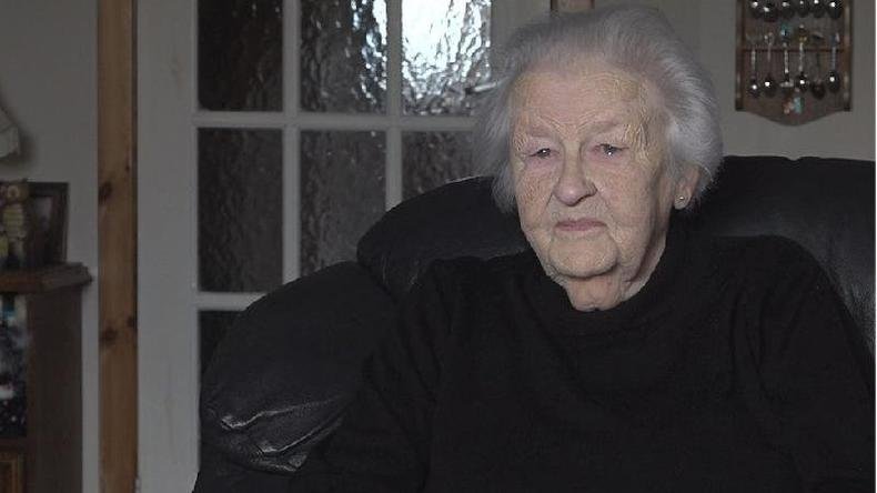 Peggy MacSween dentro de casa, sentada em poltrona, com sorriso contido