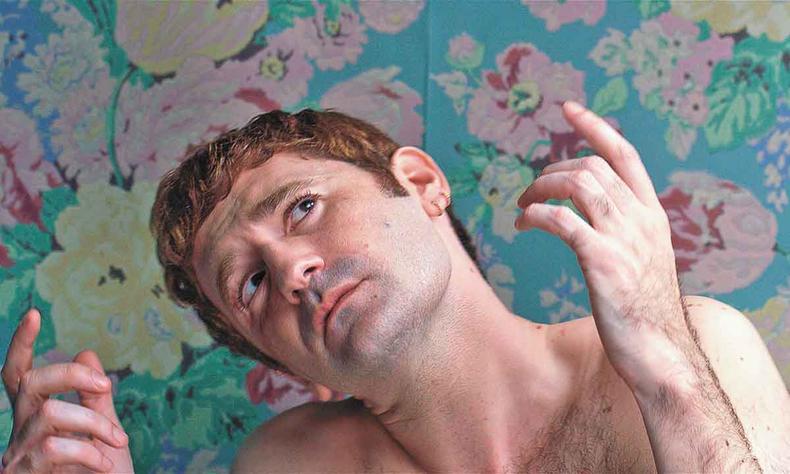 Sem camisa em ambiente com papel de parede floral, ator Nicolas Maury olha para cima e ergue as mos em cena de garoto chiffon