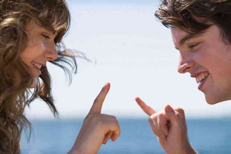 Jovem casal sorri e ergue os dedos, conversando na linguagem dos sinais, tendo o mar ao fundo em cena do filme No ritmo do coração 