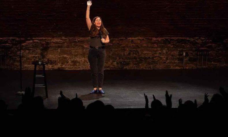 A atriz Alison Leiby com o bra�o levantado, no palco, fala de frente para a plateia