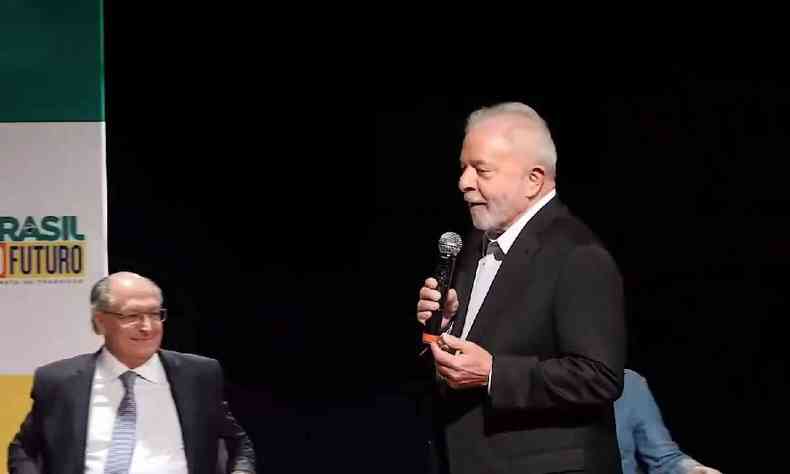 Lula em p com um microfone na mo e Geraldo Alckmin sentado em uma cadeira durante evento