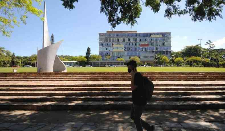 Foto do prdio da reitoria da UFMG, com monumento ao lado. Estudante passa na frente, na sombra.