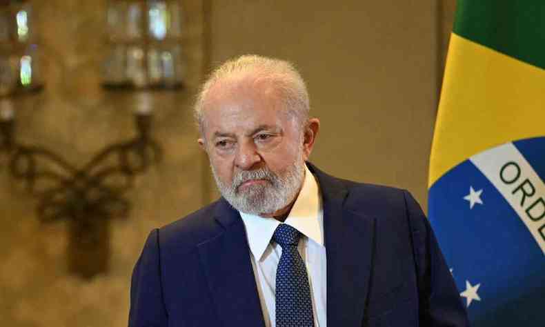 Presidente Lula, de terno escuro e expresso sria em frente a uma bandeira do Brasil