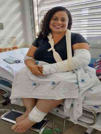 Mulher branca dos cabelos pretos e cacheados sentada na maca do hospital com tornozelo e brao enfaixados