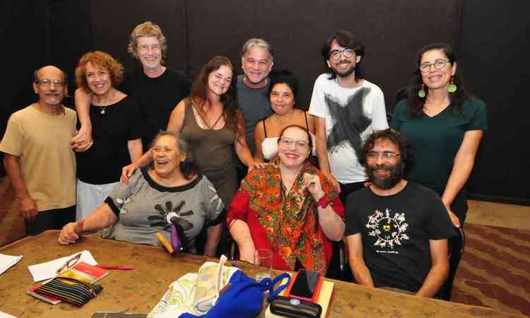 Cida Moreira sentada em frente a mesa, ao lado de Teuda Bara, e ladeada por artistas do Grupo Galpo