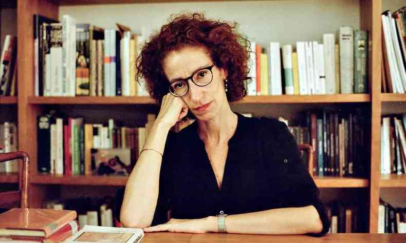 Luciana Villas Bas analisa de camisa preta e culos marrons, sentada  mesa de escritrio, com estante de livros ao fundo, apoia a mo direita no rosto