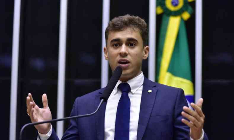 Nikolas Ferreira aparece no pulpito do congresso discursando em terno azul com a bandeira do Brasil ao fundo