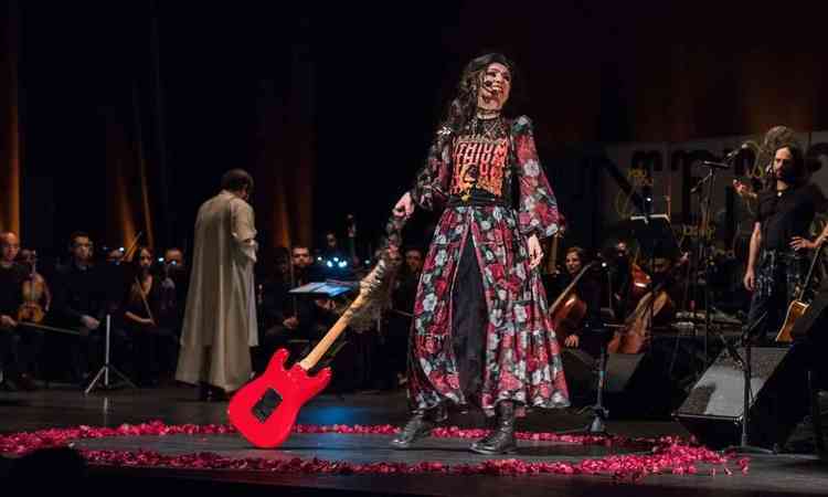 Atriz Dalila, usando vestido estampado e botas, segura guitarra durante performance no concerto da Orquestra Ouro Preto
