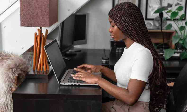 Mulher negra com cabelos longos e trançados, usando blusa branca, está sentada em uma mesa trabalhando no notebook
