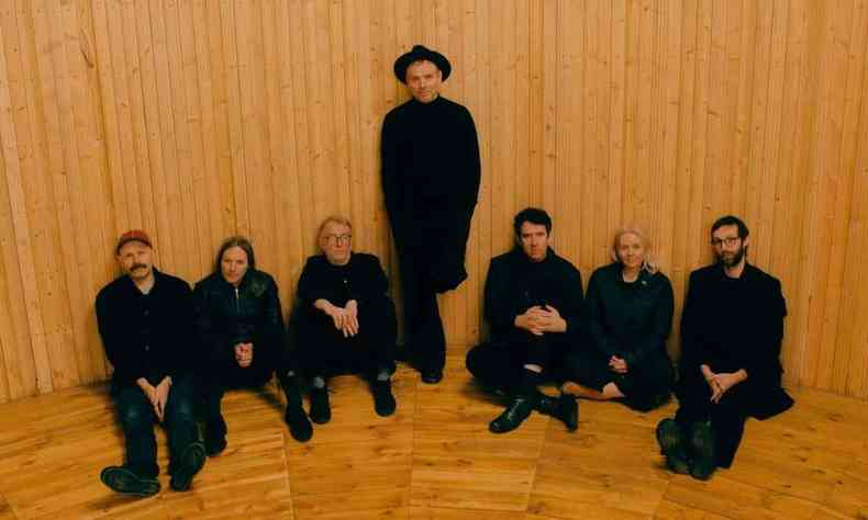 Usando chapu, o cantor Stuart Murdoch est de p, ao centro, tendo  direita e  esquerda msicos da banda Belle and Sebastian sentados no cho. Todos usam roupas pretas