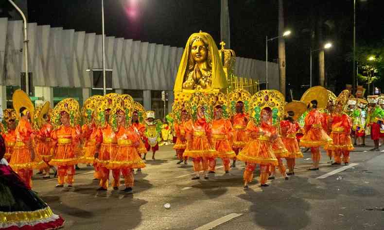 Pessoas vestidas de laranja com adereos na cabea dourados, em desfile de escola de samba em 2023. Ao fundo, santa dourada  carregada