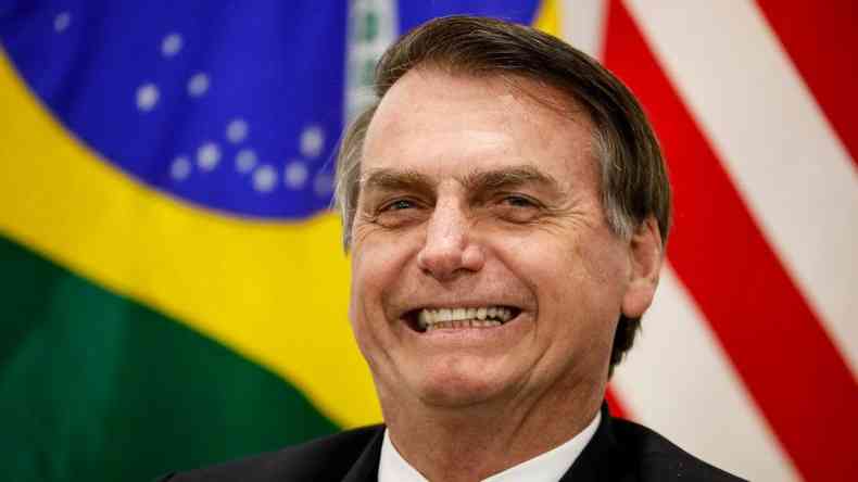 Bolsonaro sorrindo em frente a uma bandeira do Brasil