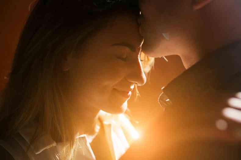 Homem com camisa escura beija testa de mulher, apesentando um semblante feliz, demonstrando um momento de afetividade e cumplicidade com uma luz ao fundo que passa entre os dois 