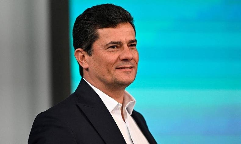 Moro em um fundo azul claro usando um terno preto, sem gravata, olhando para a direita