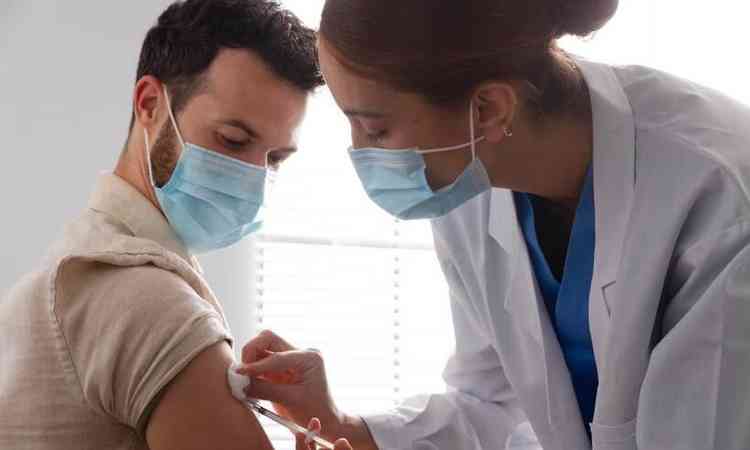 Mulher aplicando vacina no brao direito de um homem; ambos esto usando mscara cirrgica  