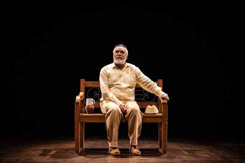 vestindo camisa e cala em tons claros, o ator Gilson de Barros, sentado em banco de madeira, com chapu ao lado, em cena de 'Riobaldo' 