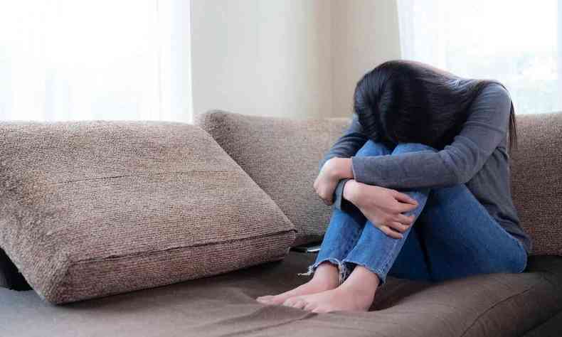 Garota sentada no sof em posio de tristeza, com a cabea baixa e os braos abraando os joelhos