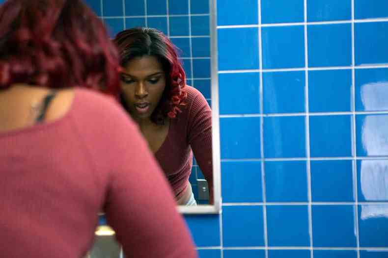 Mulher trans negra de cabelos cacheados e mechas vermelhas est em um banheiro apoiando suas mos na pia com expresso cansada