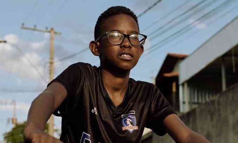 Foto do ator Ccero Lucas, adolescente negro, usando culos e uma camiseta de time de futebol enquanto anda de bicicleta.