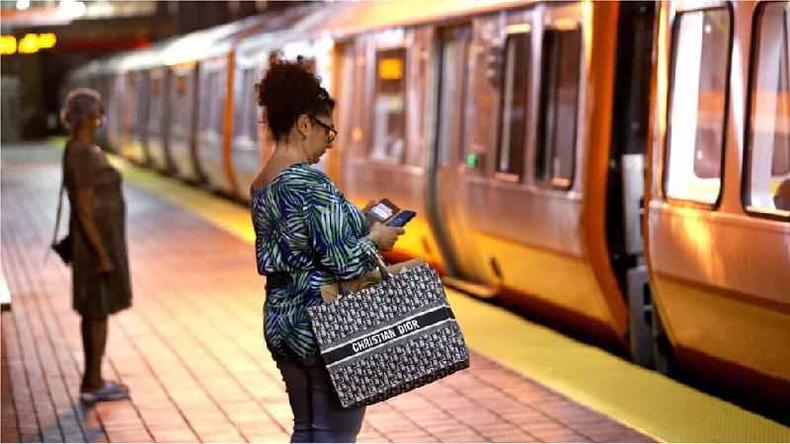 Fotografia colorida mostra mulher olhando o celular em uma estao de metro em frente a um vago