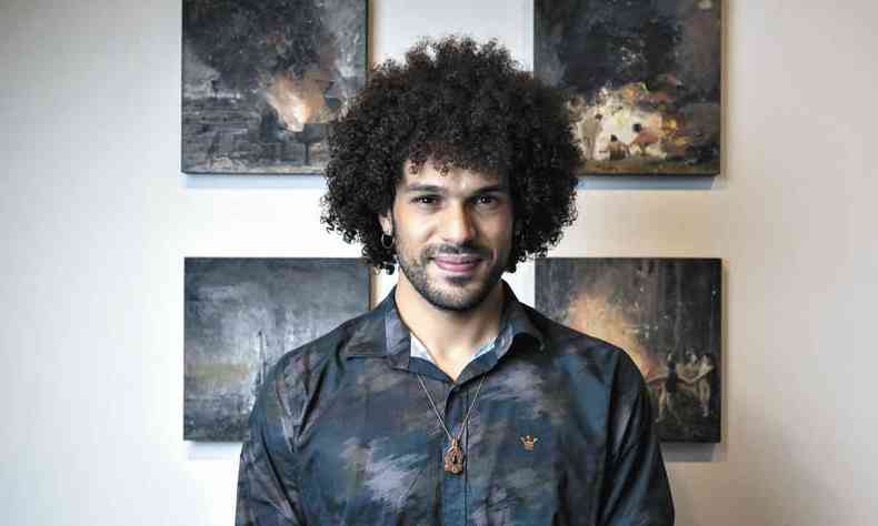 O artista Mateus Moreira sorri para a câmera, com pinturas na parede ao fundo