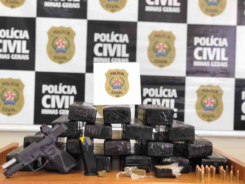Dezoito tabletes de maconha em embalagem plsticas na cor preta expostos em cima de uma mesa. Ao fundo, aparece um banner da Polcia Civil. 