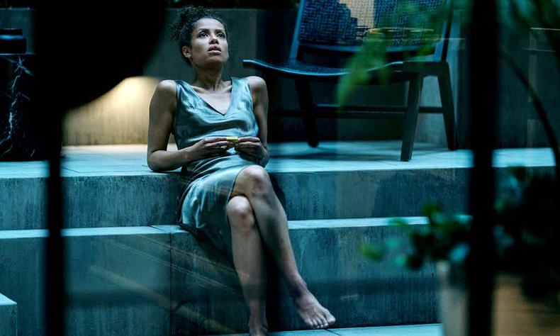 De vestido cinza, a atriz Gugu Mbatha-Raw, sentada em escada, olha para cima, em cena de 'Superfcie'
