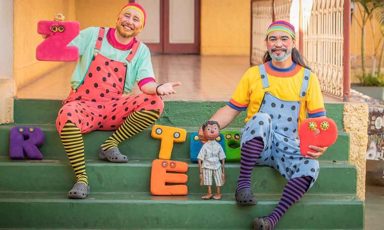 Belmiro e Adriano Borges fantasiados como palhaços estão sentados em escadas de uma varanda, segurando boneco e letras coloridas feitas de almofada