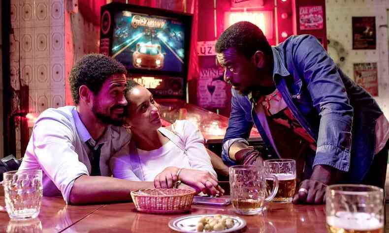 Sentados na mesa de um bar, o ator Alfred Enoch abraa a atriz Tas Arajo e ambos olham para Seu Jorge, de p, perto deles, em cena de medida provisria 