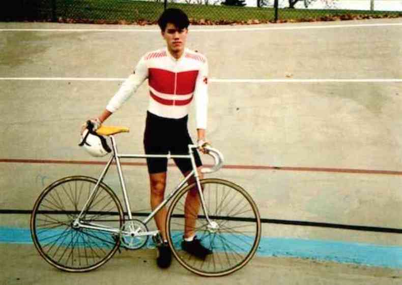 Tom Justice, um jovem magro e atlético de olhos puxados e cabelo preto, com roupas de ciclismo, ao lado de sua bicicleta