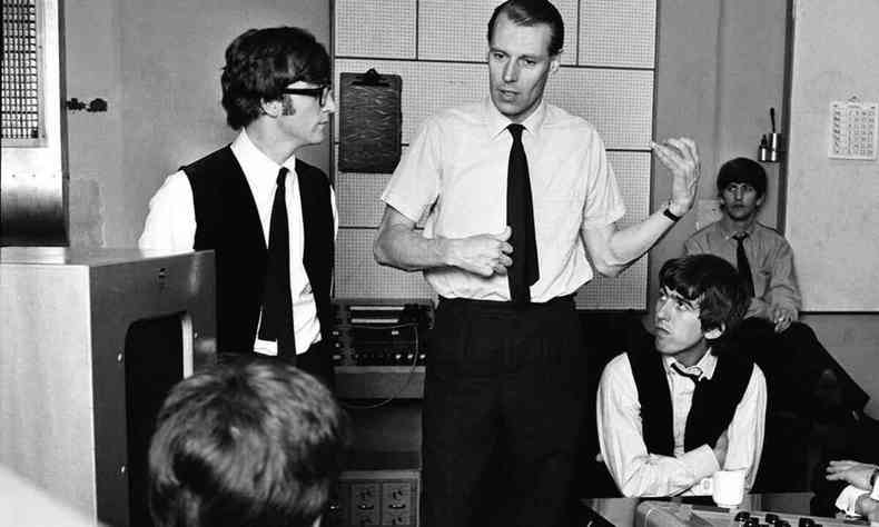 John Lennon, de culos, est em p ao lado do produtor George Martin, que faz gesto com as mos como se tocasse guitarra. George Harrison est ao lado dele, sentado, e Ringo Starr est ao fundo, sentado num banco 