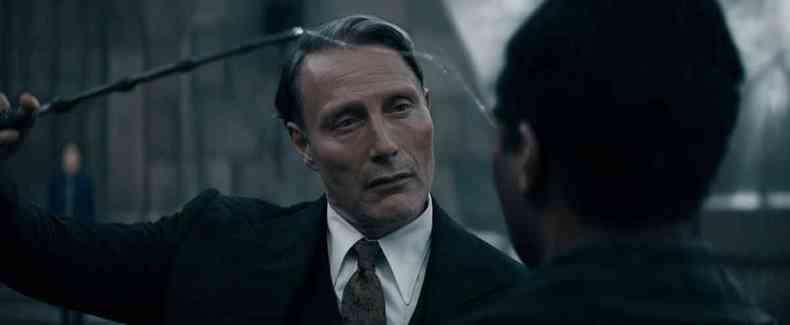 vestido de terno preto, o ator Mads Mikkelsen insere anzol da orelha de um homem em cena de o segredo de dumbledore 