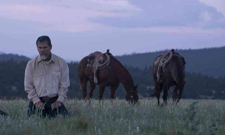 O ator Josh Brolin, vestido como caubi, est ajoelhado na grama, olhando preocupado para baixo. Ao fundo esto dois cavalos