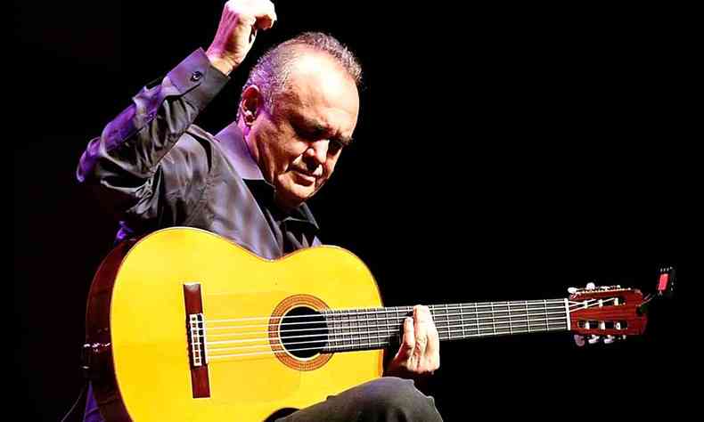De calça e camisa pretas, sentado num banquinho com seu violão, Juarez Moreira ergue a mão direita 