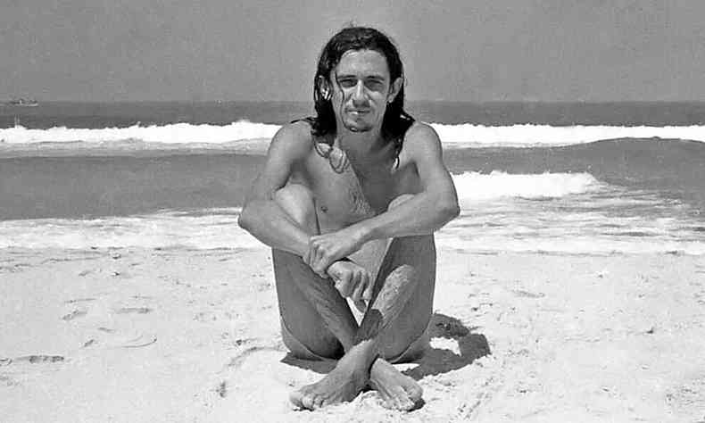 Torquato Neto, sentado na areia, de costas para o mar, com pernas e braços cruzados