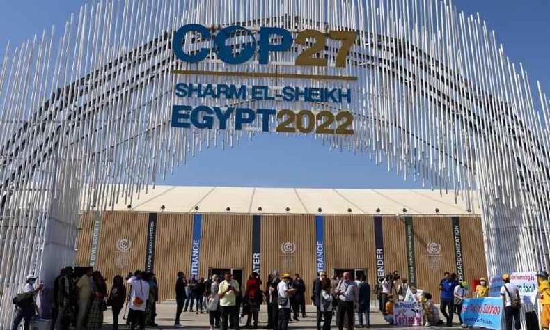 Tópico da COP-27 é demarcação de terras indígenas como forma de barrar crise climática(foto: Joseph Eid/AFP) Imagem de um arco com o logo da COP-27. Abaixo dele, há várias pessoas andando e, atrás, há uma construção alta de paredes amarelas