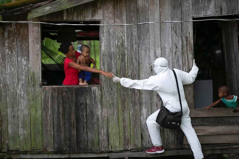 em casebre de madeira, mulher segura beb na janela e estende o brao na direo de agente de sade vestido de branco e usando mscara; outro beb circula pela casa