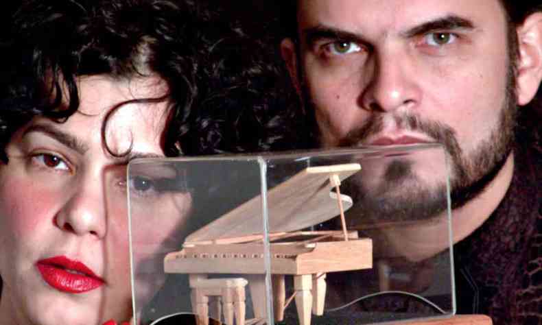 Com a miniatura de um piano na frente do rosto, a cantora Sylvia Klein e o pianista Wagner Santos olham para a cmera 