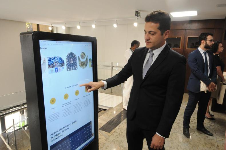  possvel ver um homem branco de terno apontando para uma tela grande onde  possvel ver o sistema Somdar aberto. Ele aponta para frase 'No sondar voc pode'