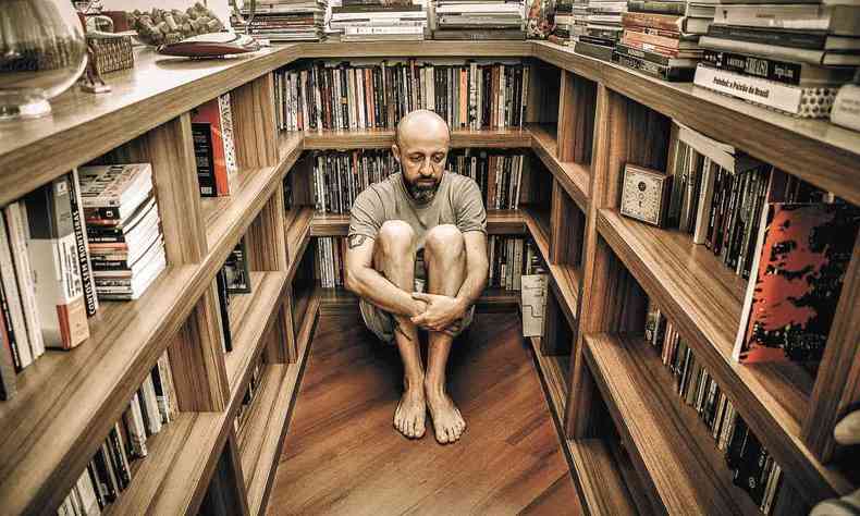 Fabrício Carpinejar sentado no chão numa biblioteca 360 graus, cabisbaixo e de bermuda