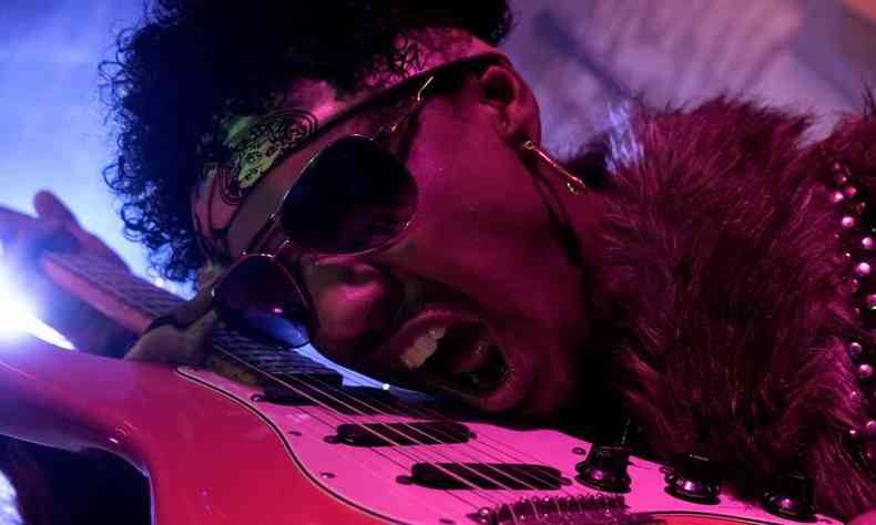 Músico Azizi, de óculos escuro e bandana colorida na cabeça, encosta o rosto na guitarra