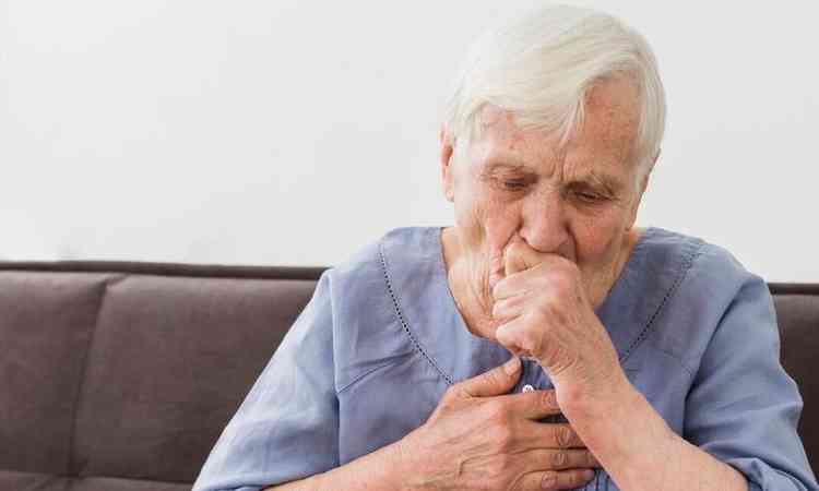 Um idoso tossindo, com uma mão cobrindo a boca e a outra na região do coração. 