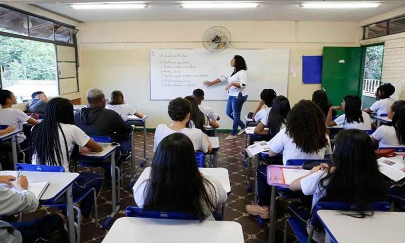 uma sala de aula cheia com a professora ensinando ao fundo; quadro tem umas escritas
