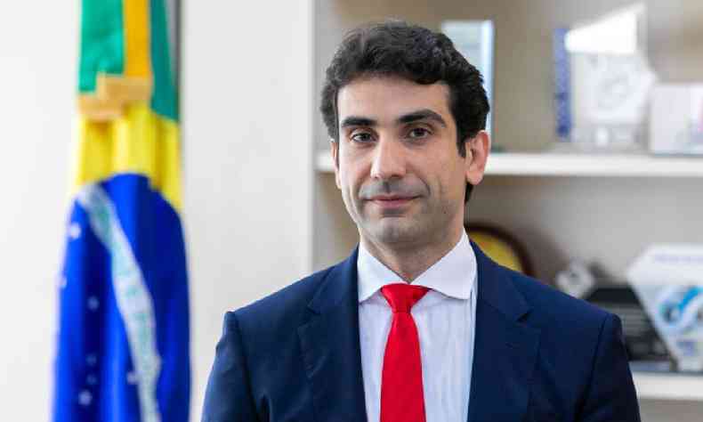 Gabriel Galpolo de terno azul marinho, camisa branca e gravata vermelha com a bandeira do Brasil ao fundo